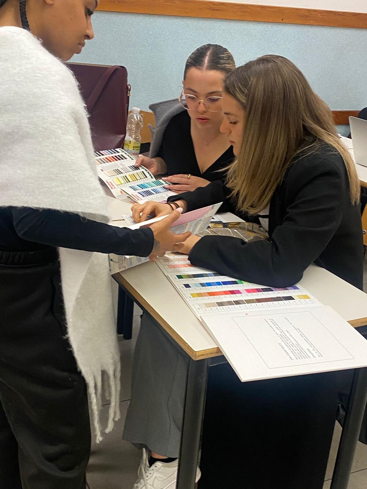 Tre studentesse confrontano cartelle colore e campioni di tessuto su un banco durante una lezione.
