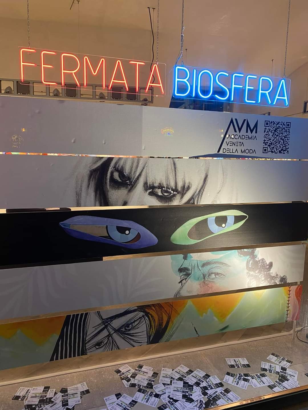 La parete della mostra con i pannelli dipinti di occhi e volti, sotto le insegne al neon «Fermata» e «Biosfera» e il logo dell’Accademia.